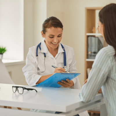 A doctor in a white coat smiles while taking notes on a clipboard during a consultation with a patient.